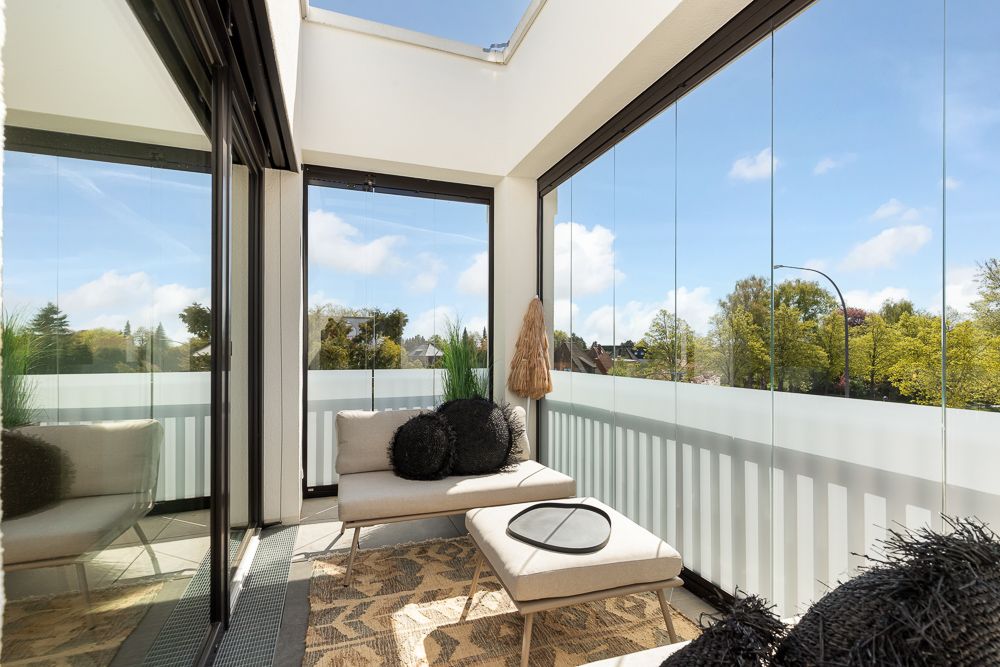 Heller Balkon mit Glaswänden, Sitzmöbeln und Kissen, Blick auf Bäume im Freien.