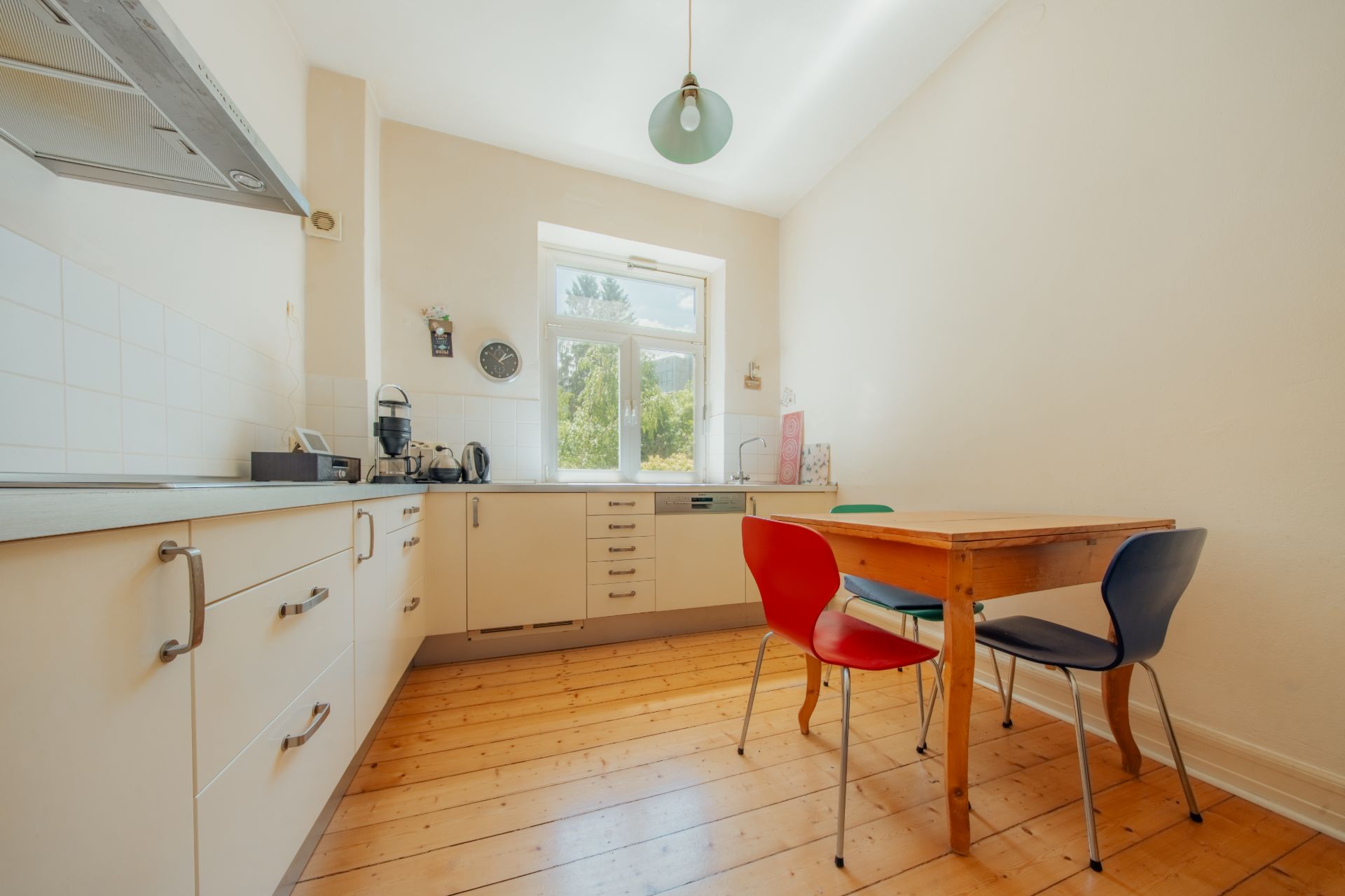 Helle Küche mit Holzboden, Esstisch und zwei bunten Stühlen vor einem Fenster.