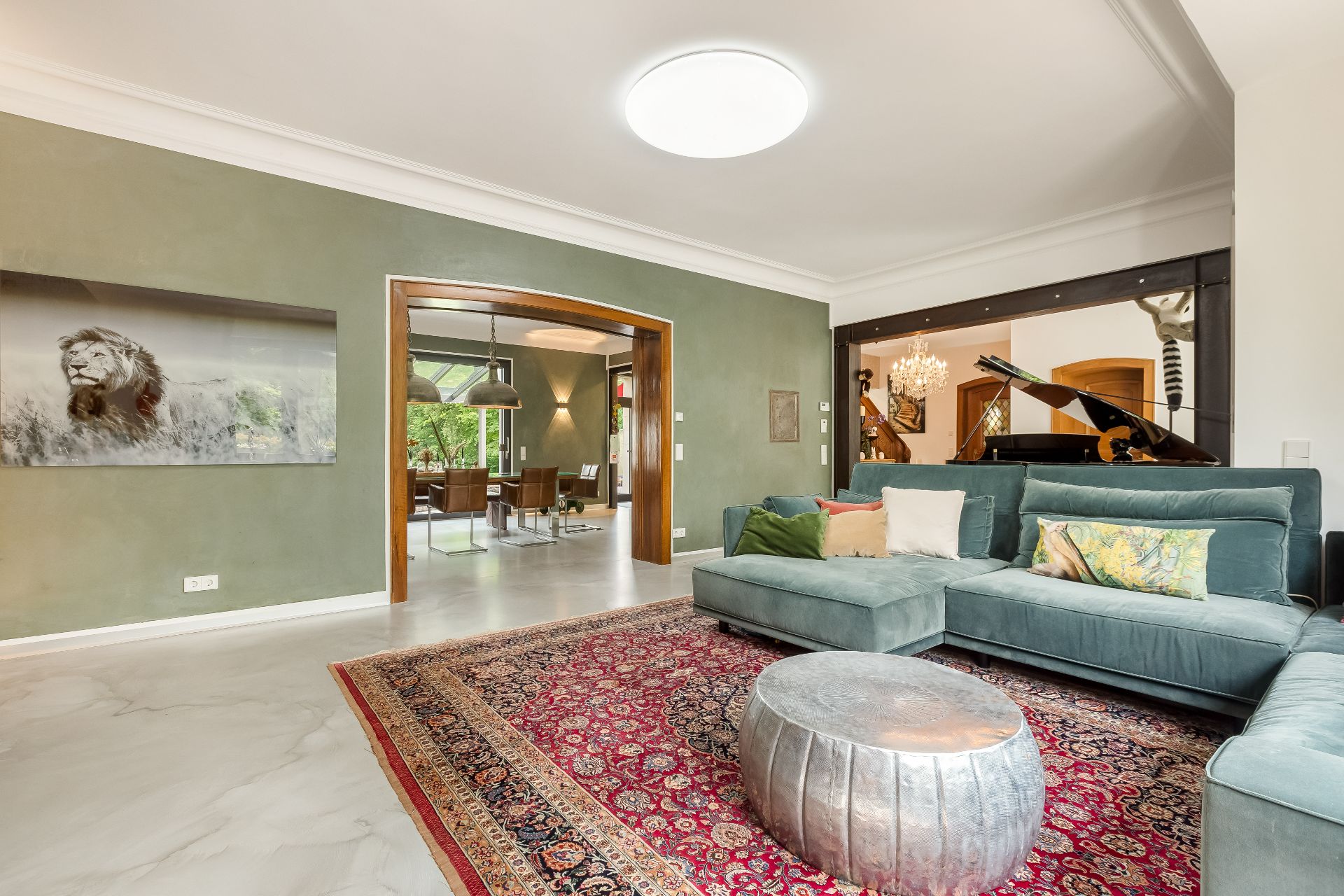 Wohnzimmer mit grünem Sofa, rotem Teppich, silbernem Tisch und großem Klavier im Hintergrund.