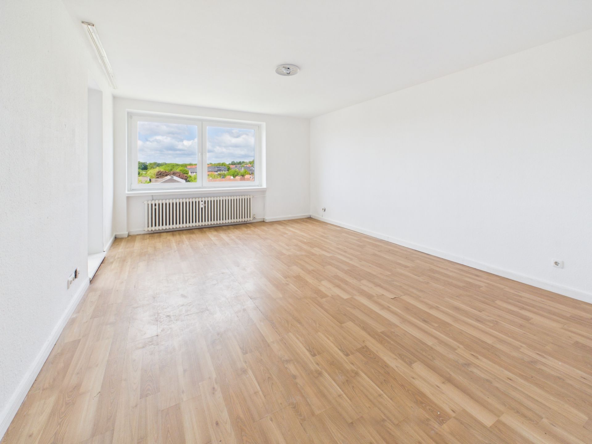 Heller, leerer Raum mit Holzfußboden und großem Fenster, durch das man nach draußen sieht.
