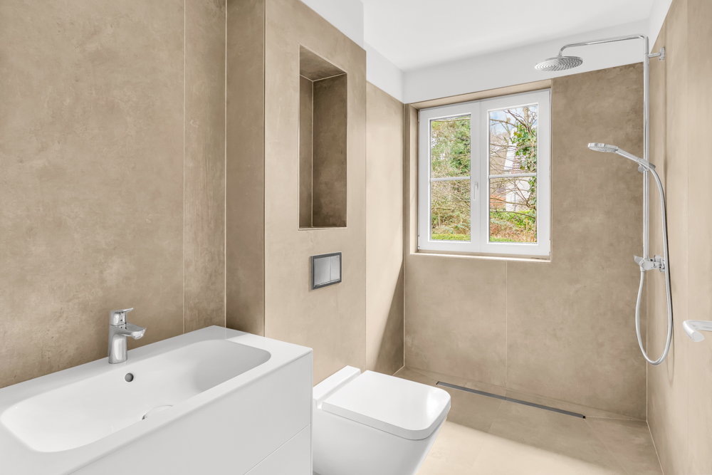 Modernes Badezimmer mit beiger Wand, Waschbecken, Toilette, offenem Duschbereich und Fenster.