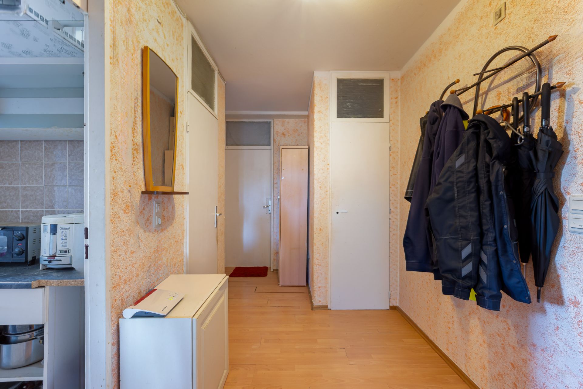 Flur mit Garderobe, Jacken und Regenschirmen an der Wand, sichtbare Küchenecke links.