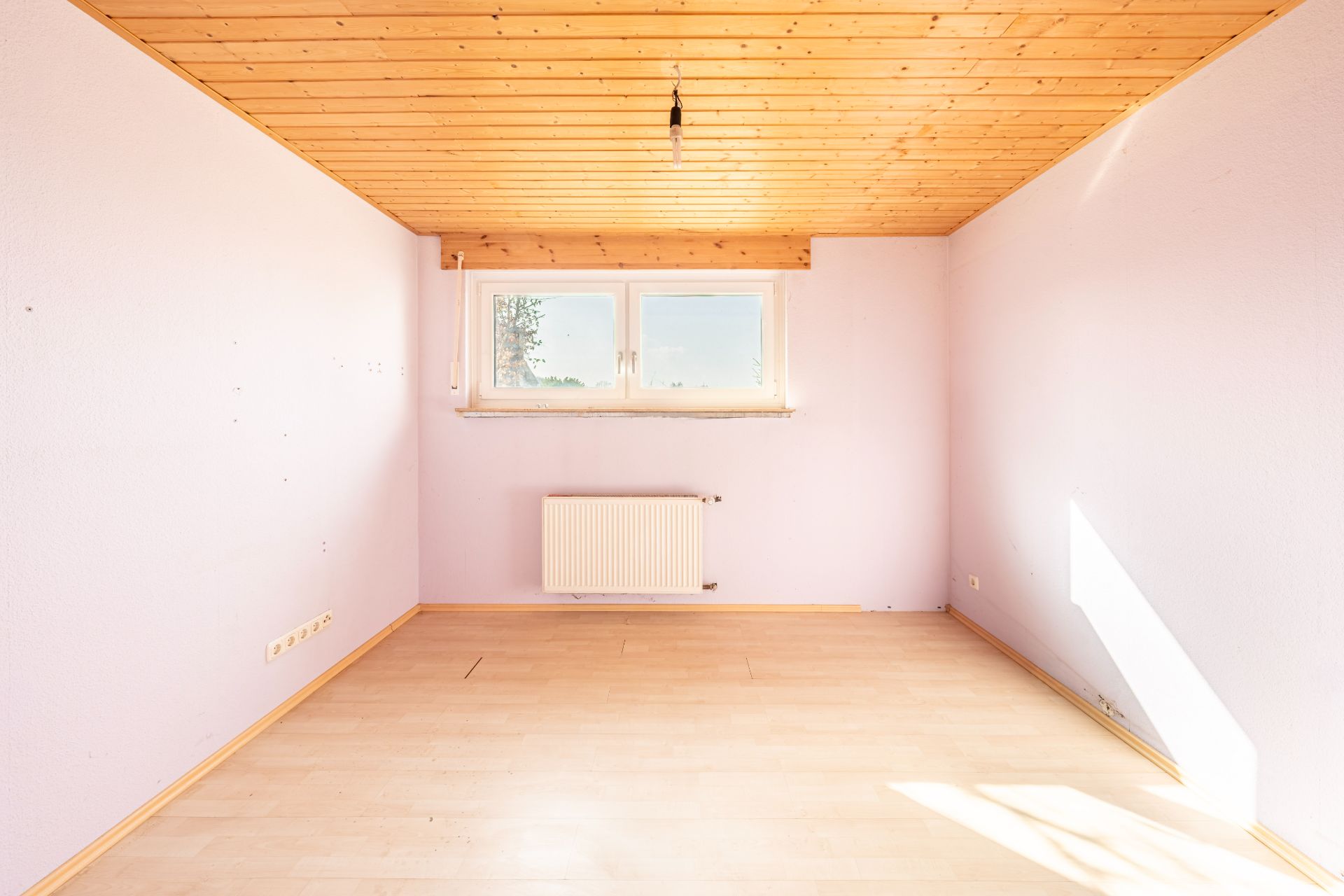 Leerer, heller Raum mit Holzdecke, Fenster, Heizkörper und hellem Laminatboden.
