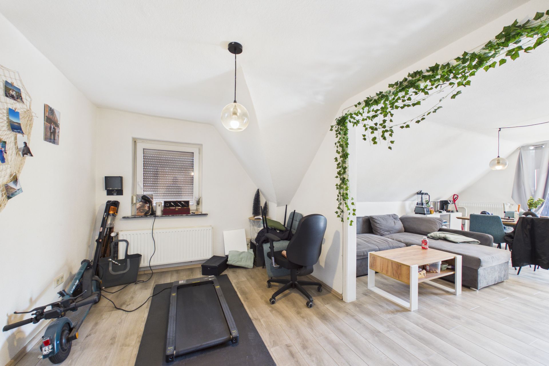 Wohnzimmer mit Schreibtisch, Sofa, Laufband und Esstisch unter Dachschrägen, dekoriert mit Zimmerpflanzen.