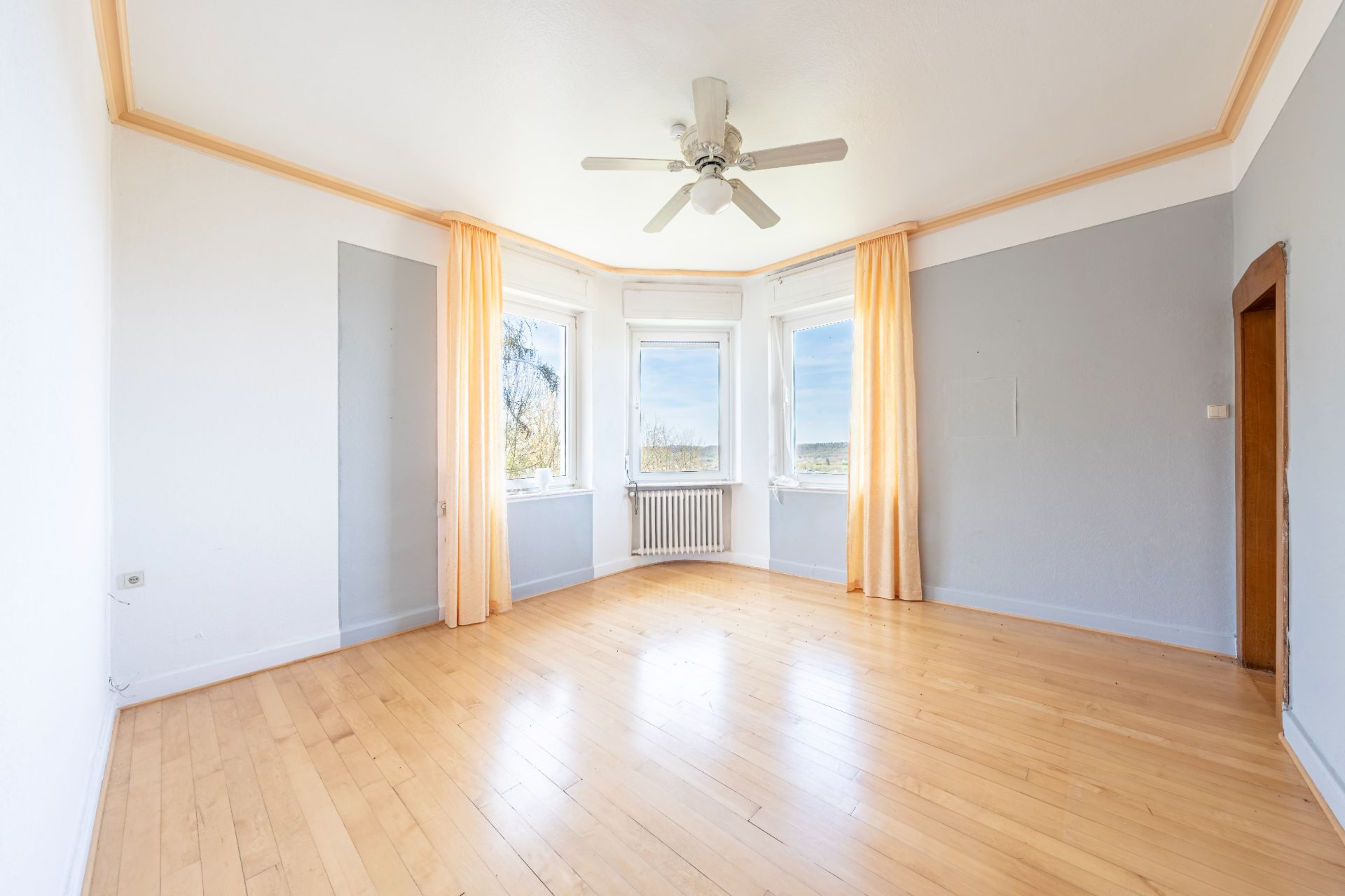 Heller, leerer Raum mit Holzfußboden, drei Fenstern, Deckenventilator und gelben Vorhängen.