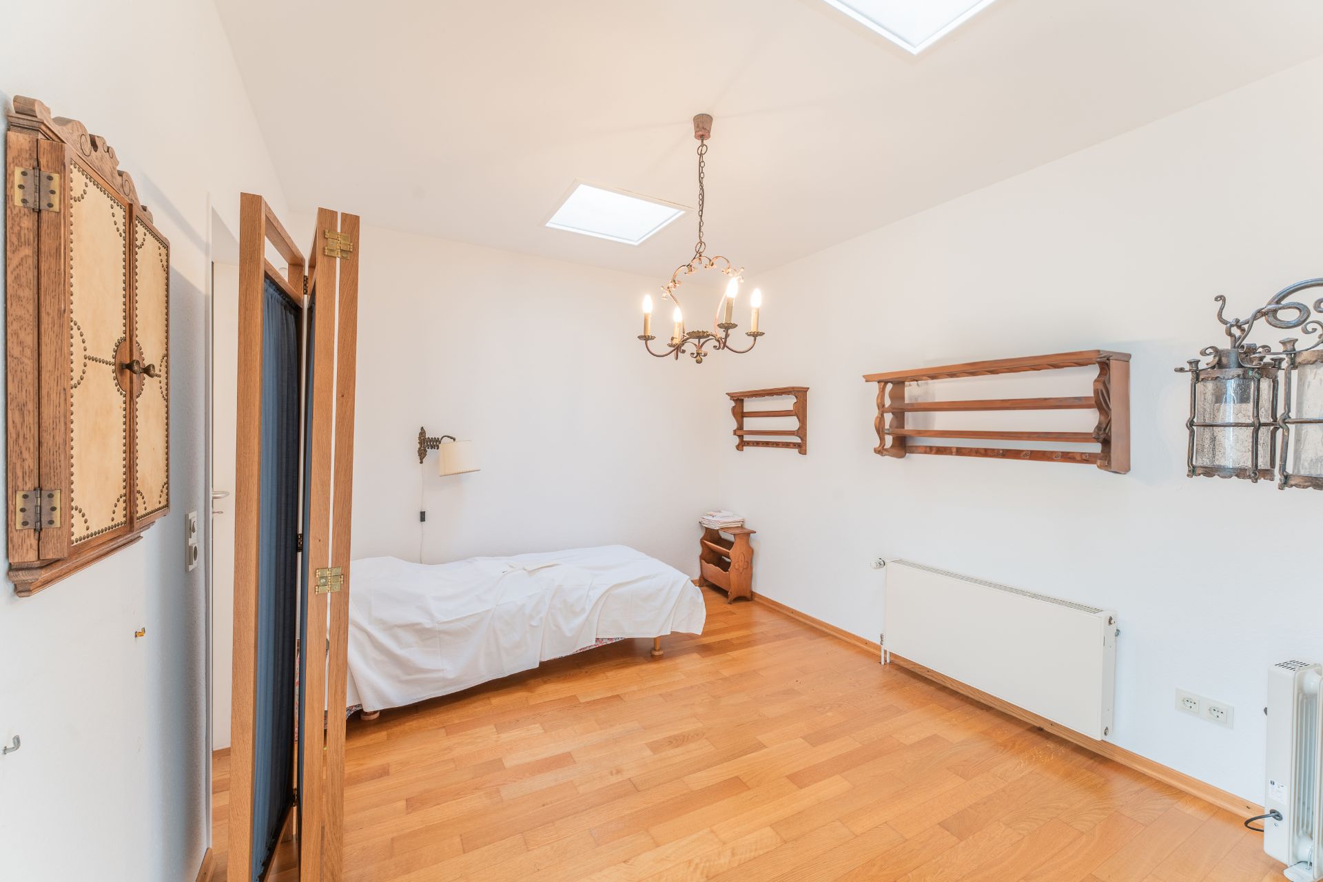 Helles Schlafzimmer mit Holzboden, Einzelbett, Kronleuchter und Holzregalen an der Wand.