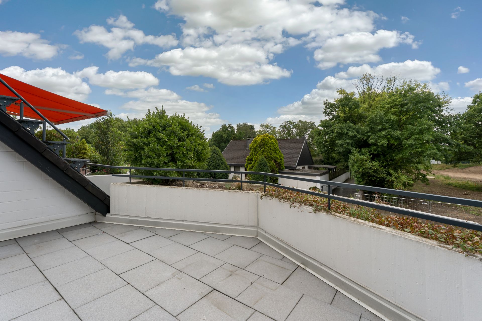 Große, geflieste Terrasse mit Geländer und Blick auf Bäume und Häuser unter bewölktem Himmel.