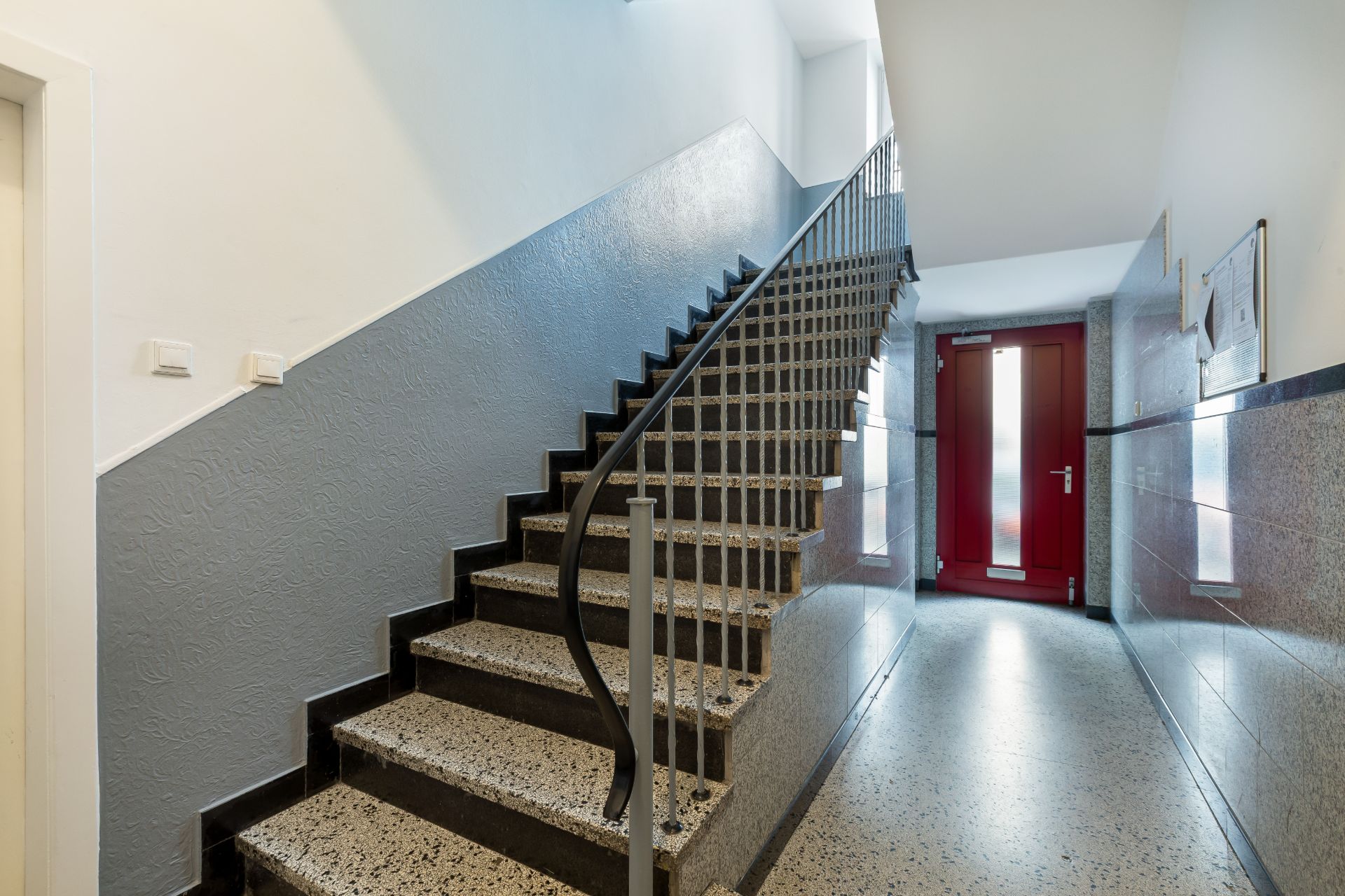 Treppenhaus mit Steintreppe, Metallgeländer und rotem Eingangstür im Hintergrund.