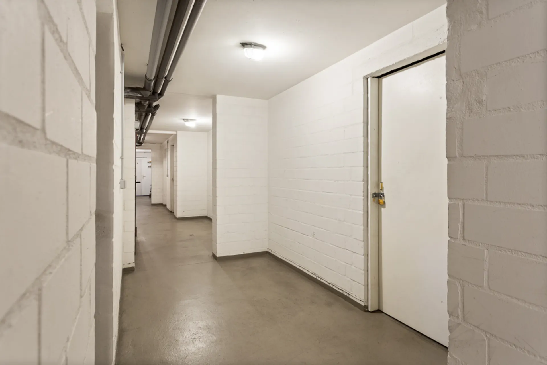 Heller Flur mit weißen Wänden, mehreren Türen und sichtbaren Rohrleitungen an der Decke.