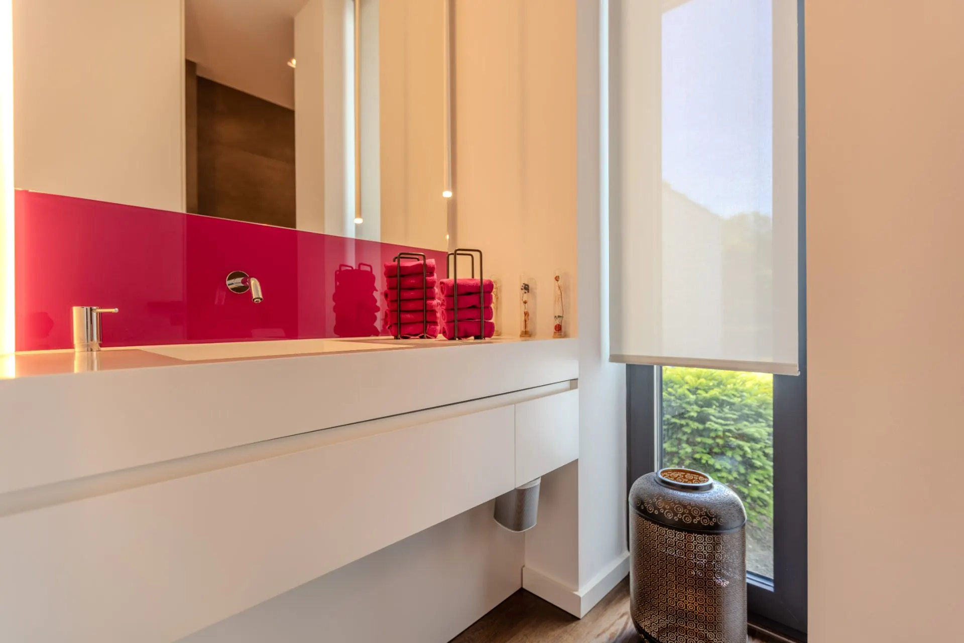 Modernes Badezimmer mit weißem Waschbecken, pinker Rückwand und großem Fenster mit Rollo.