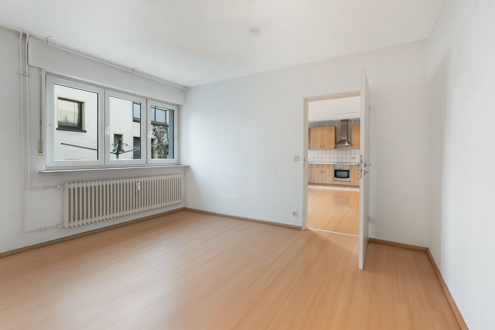 Helles, leeres Zimmer mit Laminatboden, Fenster und Blick in eine Küche mit Holzmöbeln.