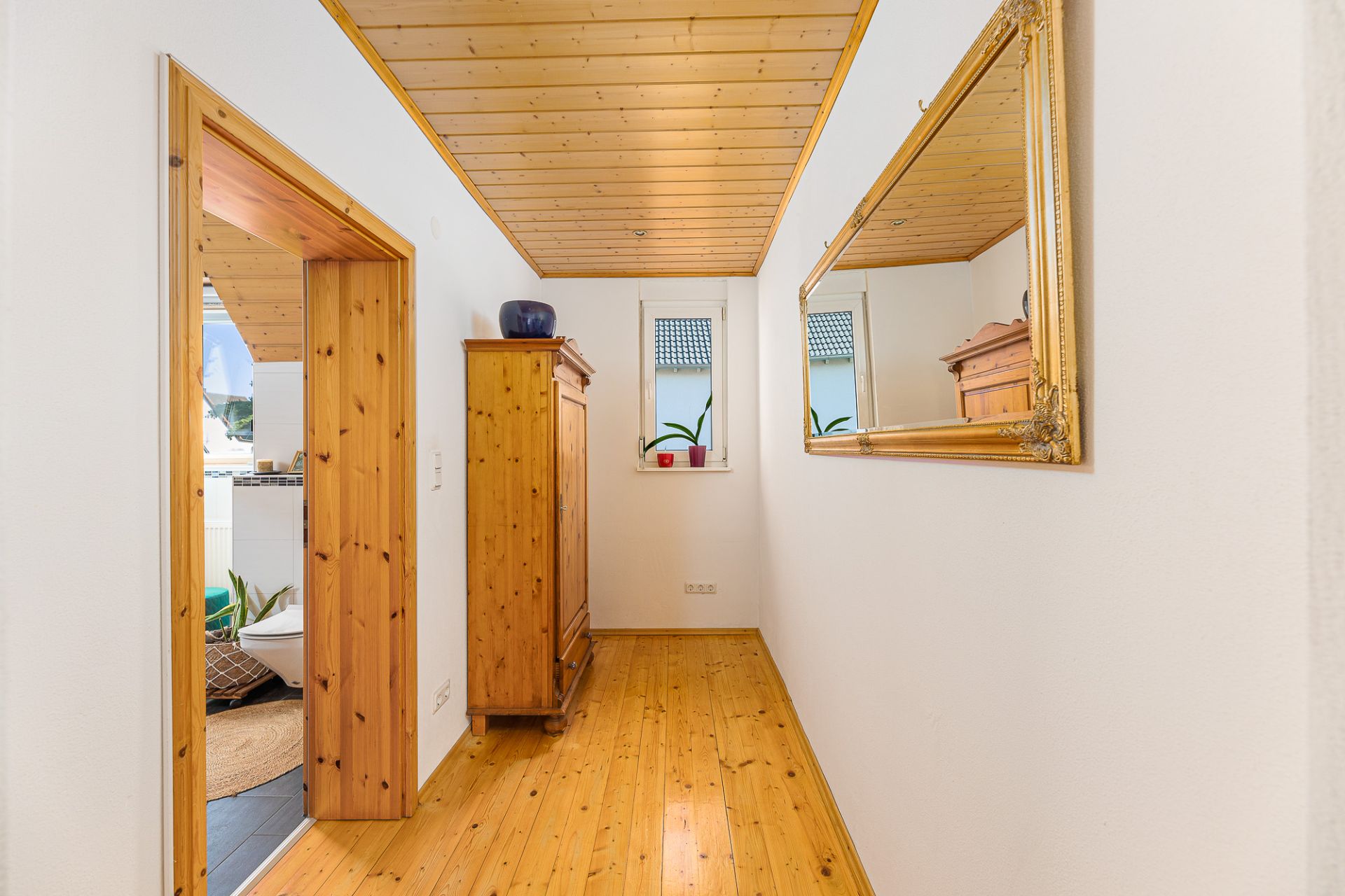 Heller Flur mit Holzdecke, Holzschrank, großem Spiegel und zwei Fenstern mit Pflanzen.