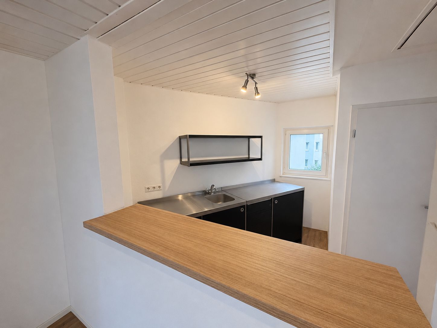 Moderne kleine Küche mit Holztheke, Spüle, Hängeregal und Fenster, weiße Wände und Decke.