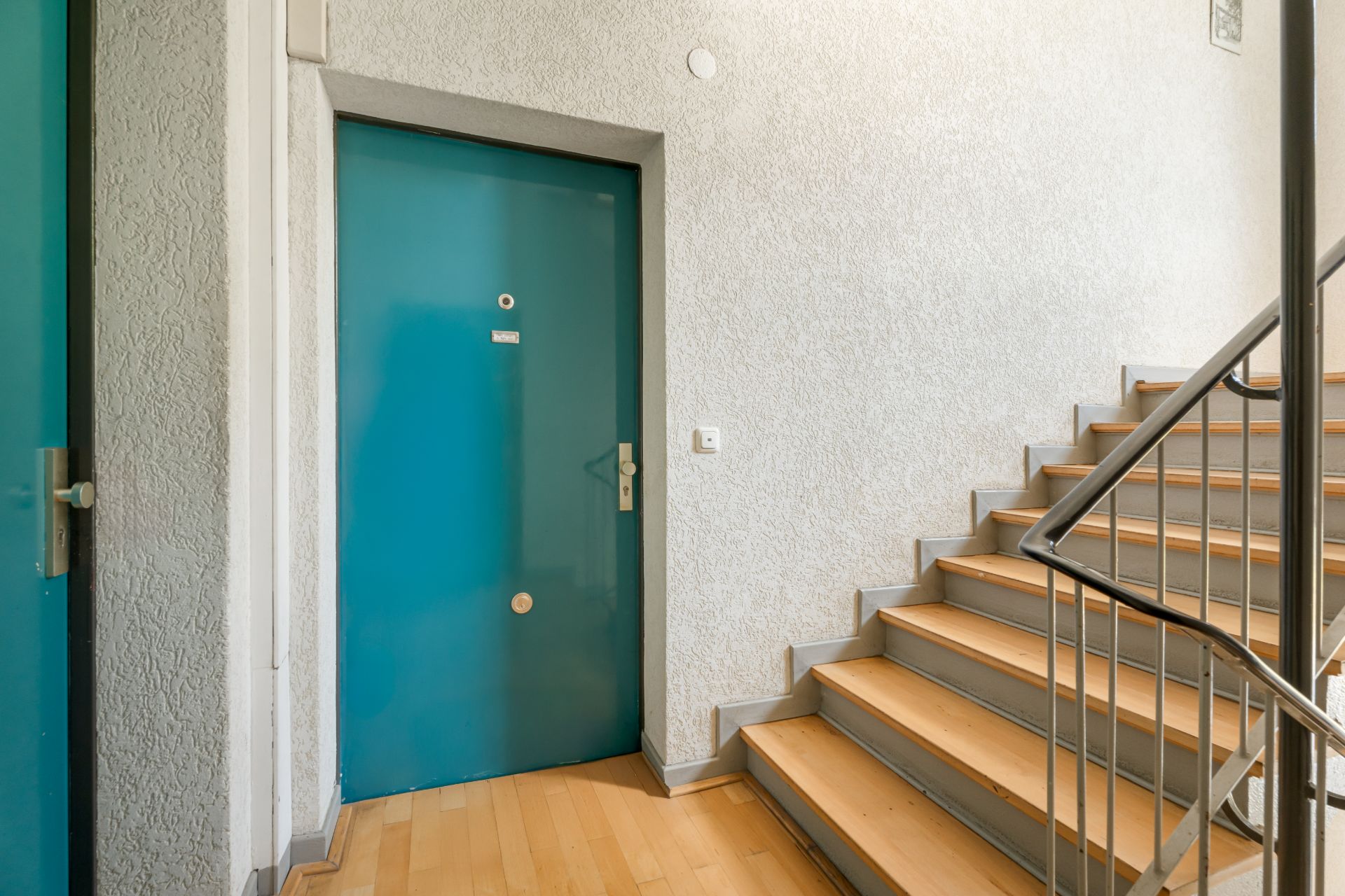 Treppenhaus mit blauen Wohnungstüren und Holztreppe mit Metallgeländer.