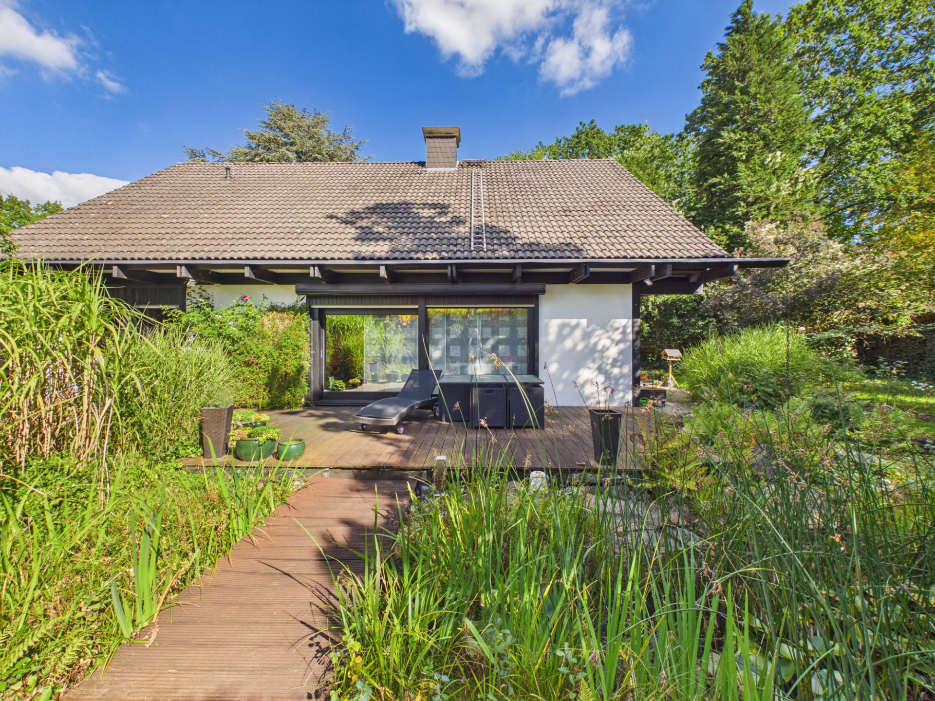 Einfamilienhaus mit Terrasse, Holzsteg und Garten, umgeben von viel Grün.