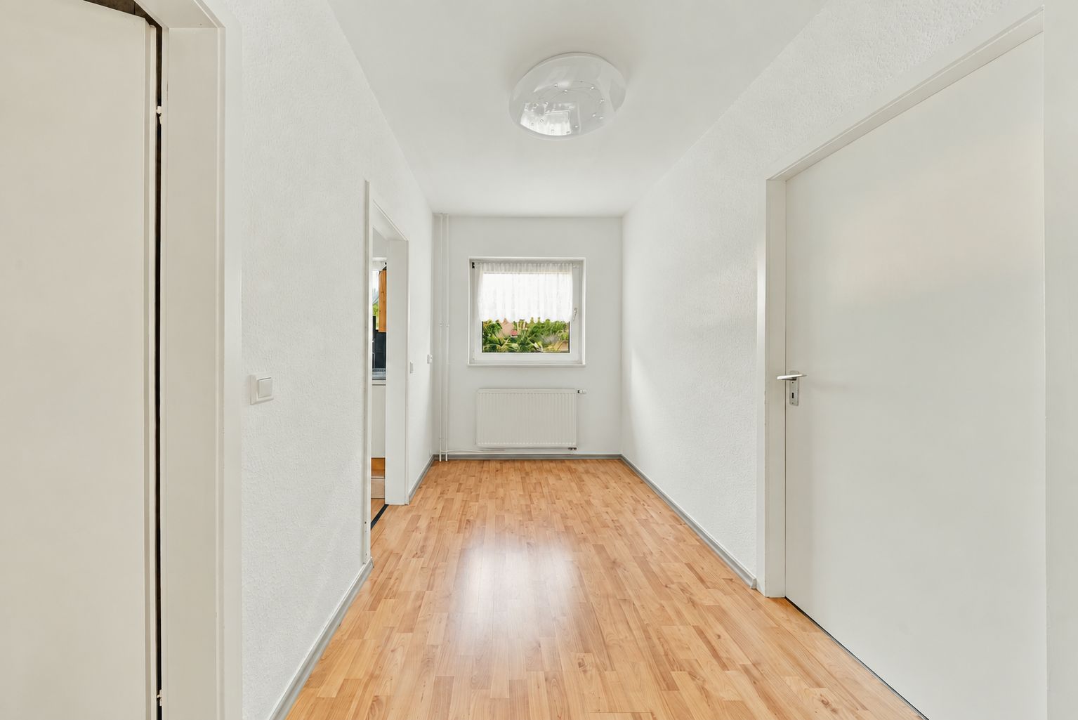 Heller Flur mit Holzfußboden, weißen Wänden und einem Fenster am Ende.