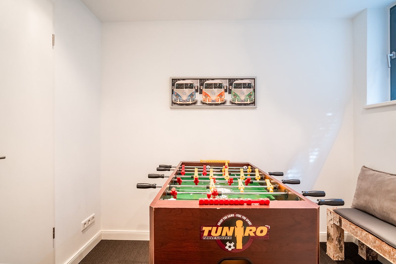 Ein Kicker steht in einem hellen Raum mit weißer Wand und kleinem Sofa.