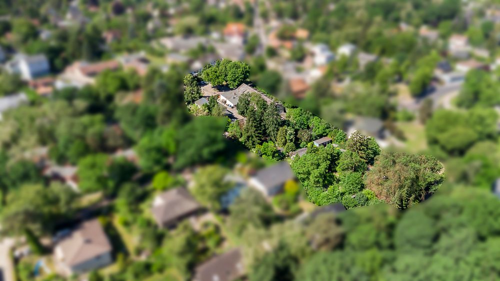 Luftbild eines Wohnhauses mit umgebenden Bäumen und Garten, unscharfe Nachbarschaft im Hintergrund.