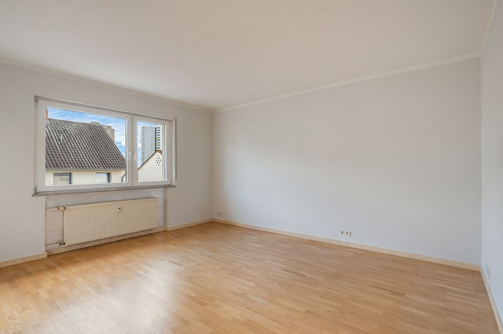 Helles, leeres Zimmer mit Holzfußboden, weißer Wand und großem Fenster.