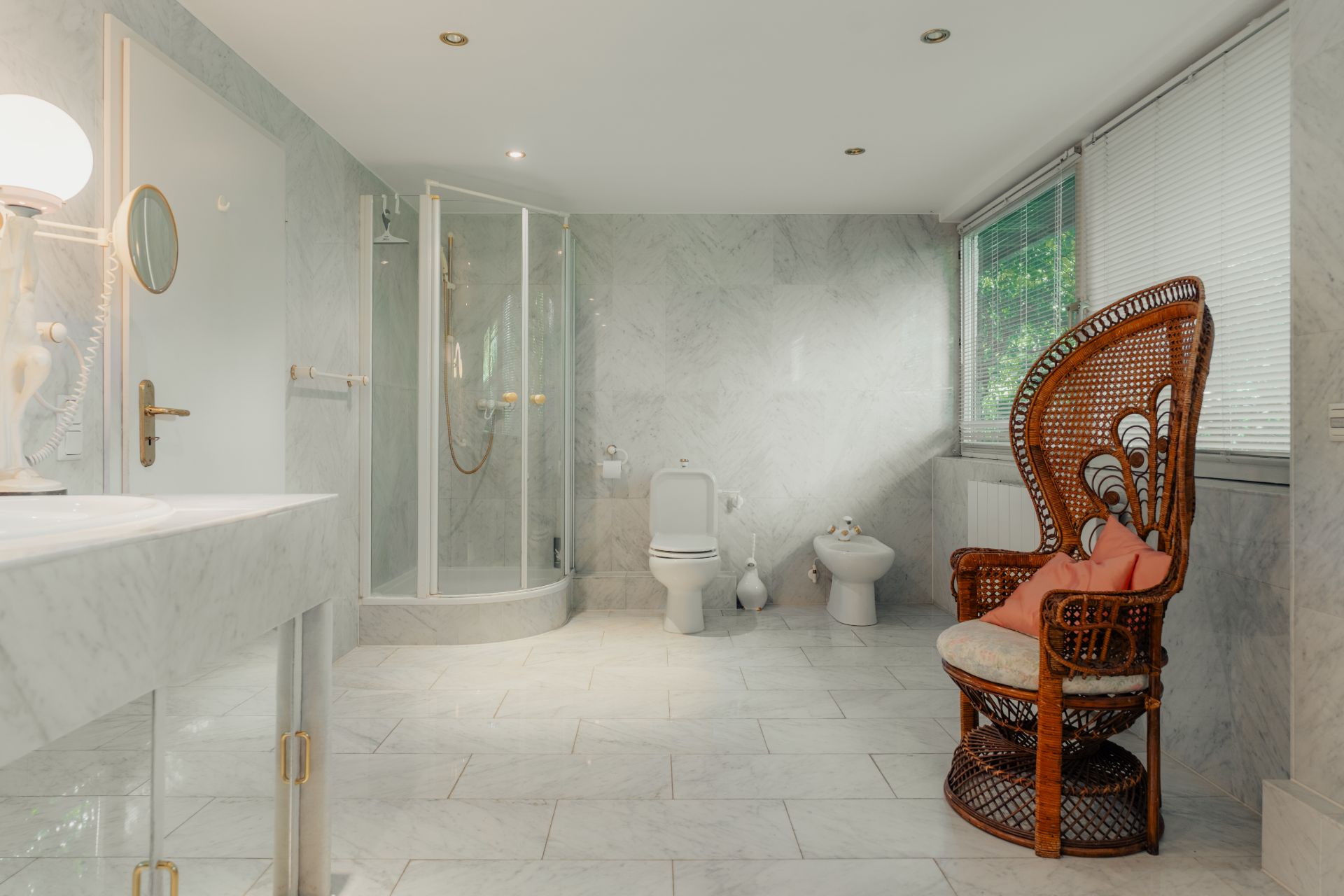 Großes Badezimmer mit Dusche, WC, Bidet, Waschtisch und Korbstuhl vor einem Fenster.