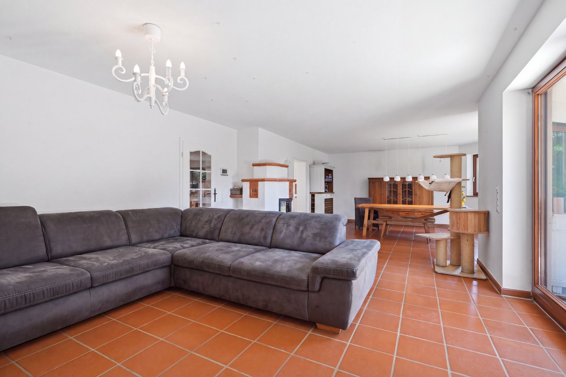 Wohnzimmer mit grauem Ecksofa, Kronleuchter und Essbereich auf Fliesenboden.