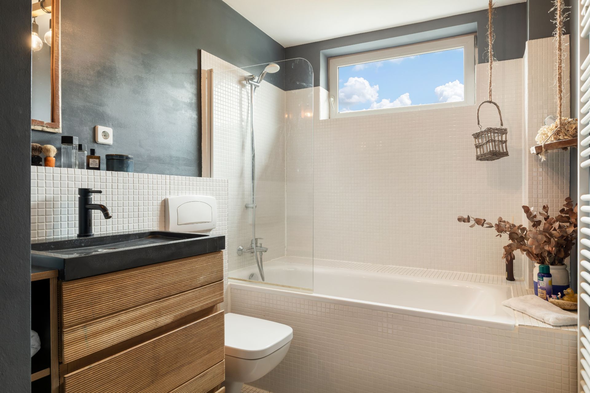 Modernes Badezimmer mit Badewanne, Waschbecken und Fenster, helle Fliesen und schwarze Armaturen.