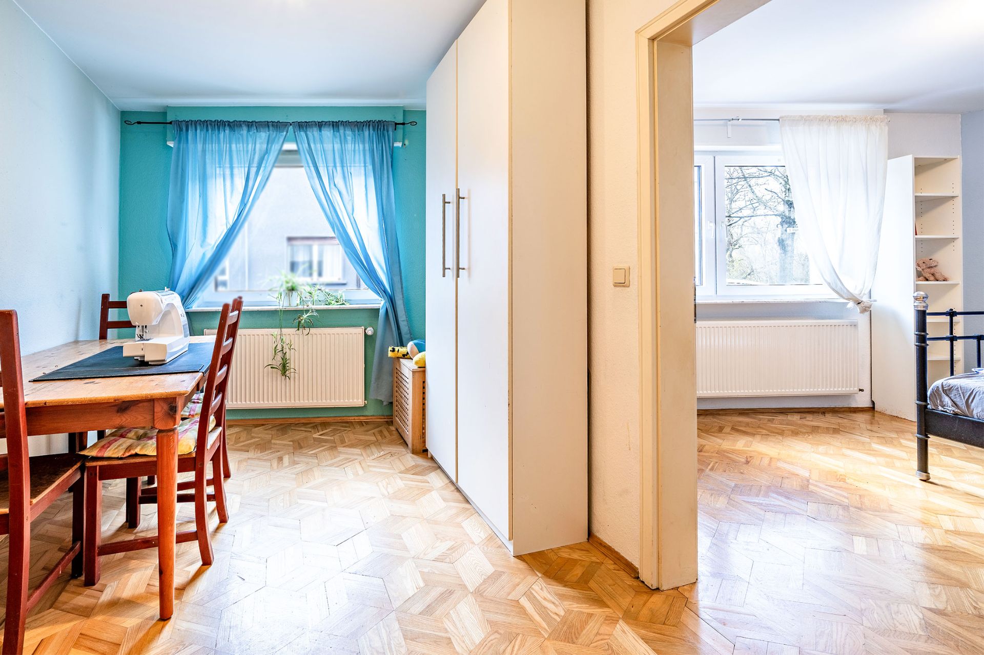 Helles Zimmer mit Holztisch, Nähmaschine, zwei Stühlen, blauem Vorhang und Blick ins Schlafzimmer.