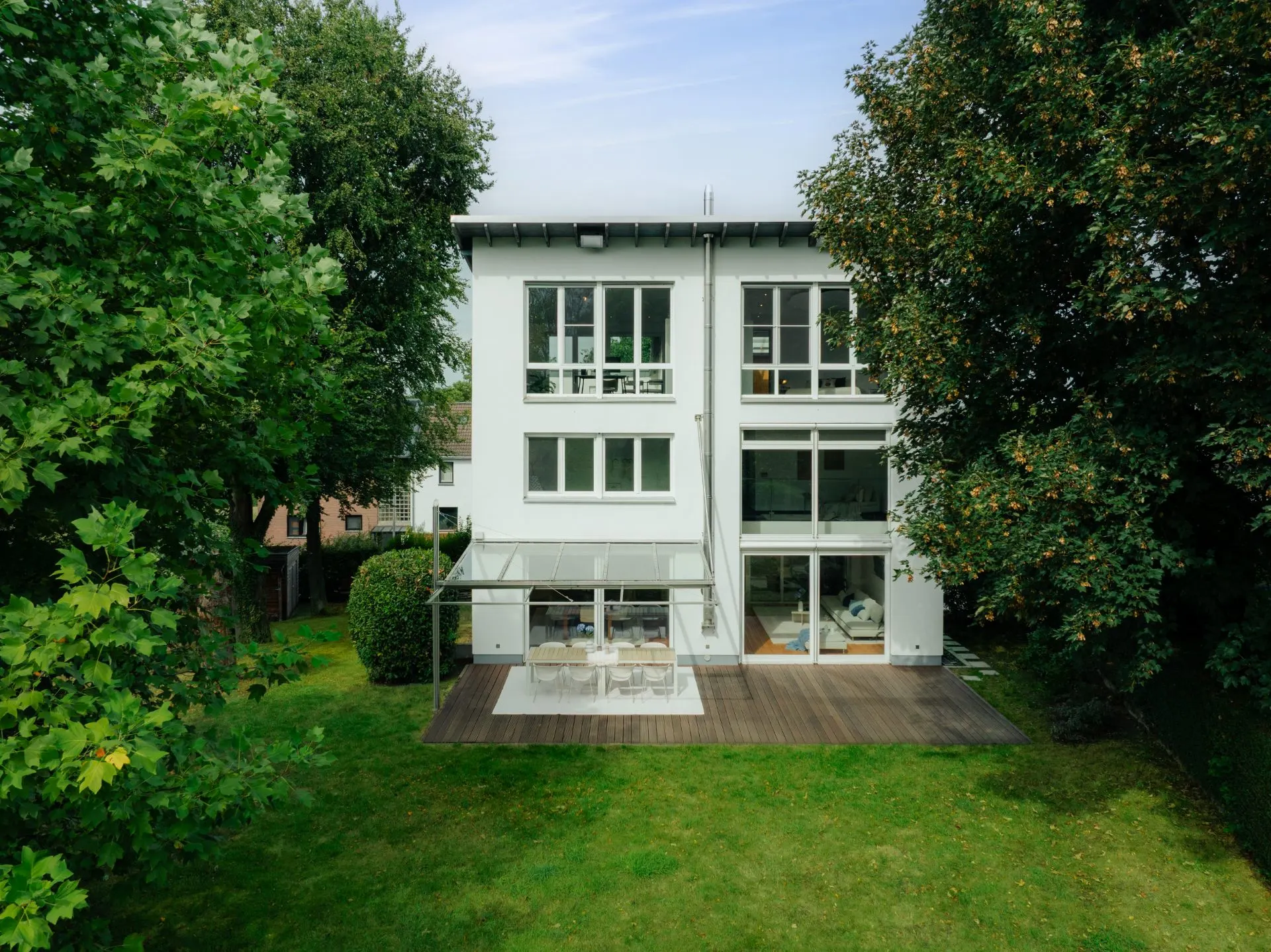 Modernes weißes Wohnhaus mit großer Terrasse und Glasüberdachung, von grünem Garten umgeben.