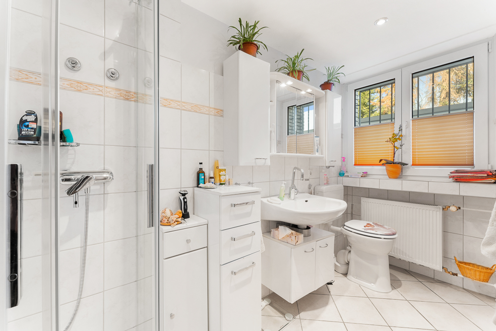 Helles Badezimmer mit Dusche, Waschbecken, Toilette, Spiegelschrank und Pflanzen auf den Schränken.