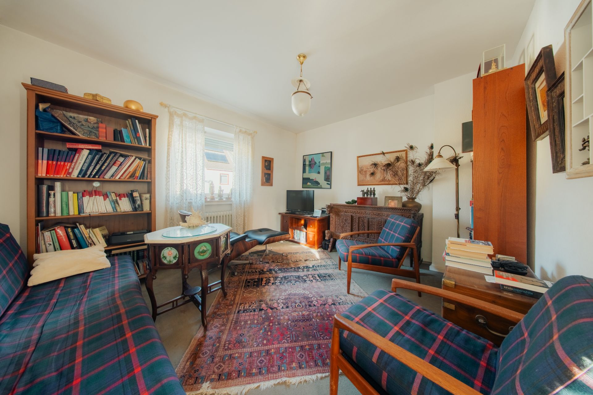 Wohnzimmer mit Teppich, Bücherregal, zwei Sesseln, Sofa und Fenster mit Vorhang.
