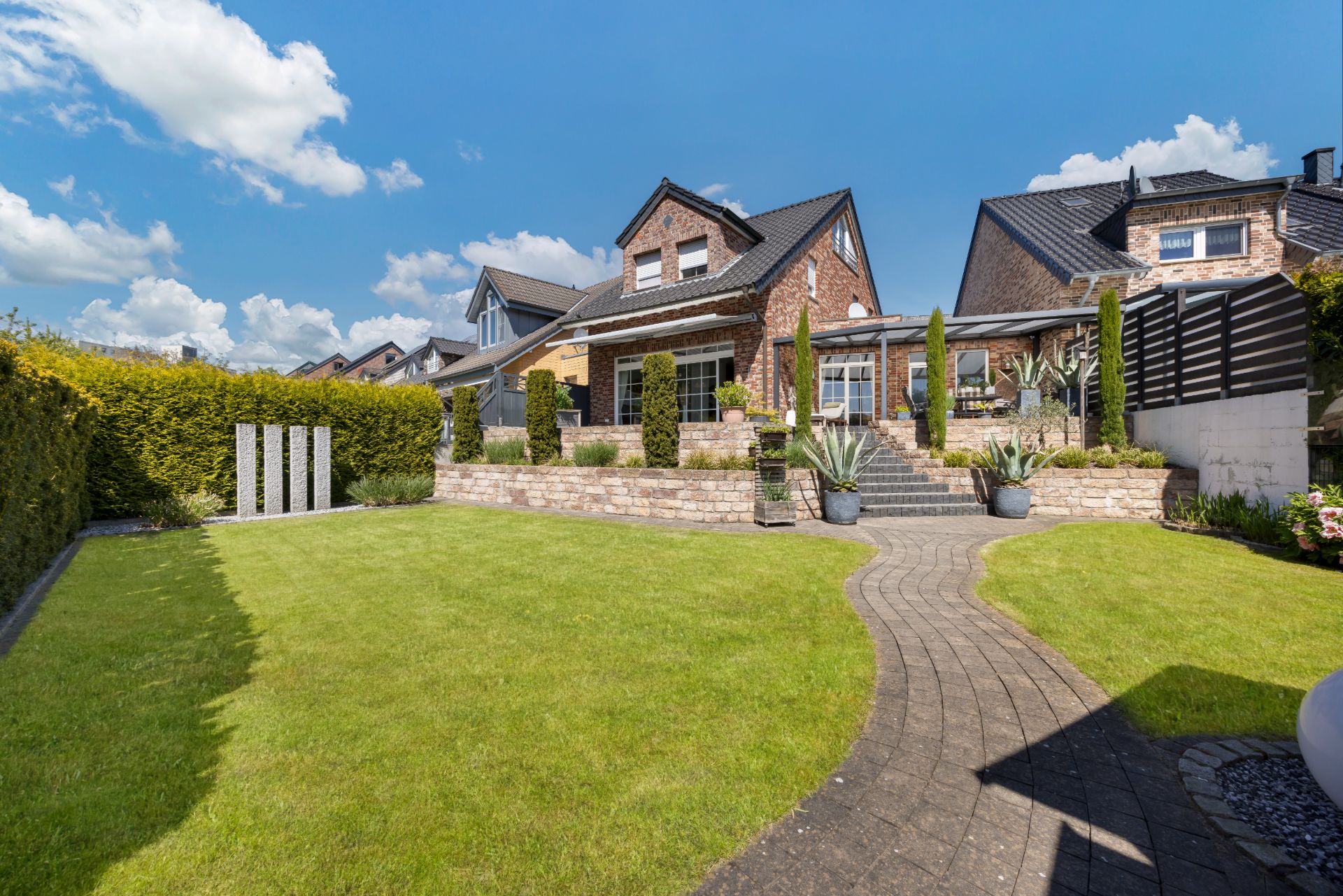 Gepflegter Garten mit Rasen, Steinweg und Backsteinhaus im Hintergrund.