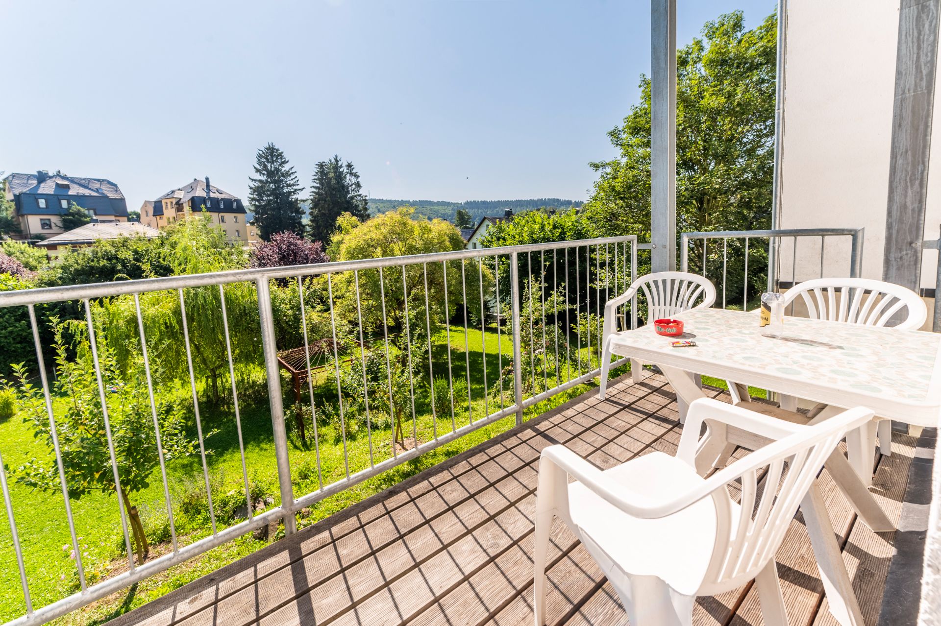 Balkon mit weißem Tisch und Stühlen, Blick auf grünen Garten und Bäume.