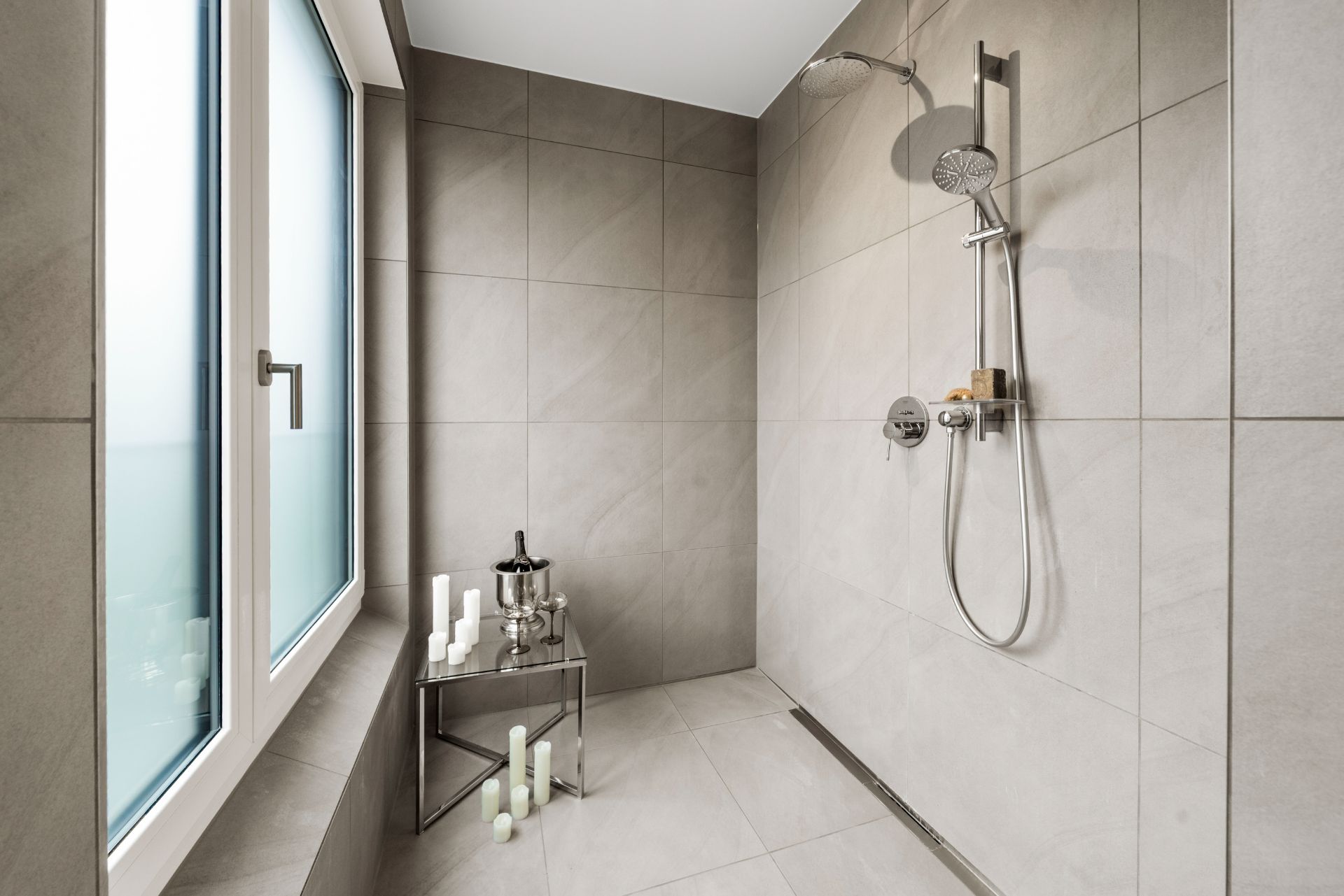 Moderne, offene Dusche mit grauen Fliesen, Fenster und kleinem Metalltisch mit Kerzen.