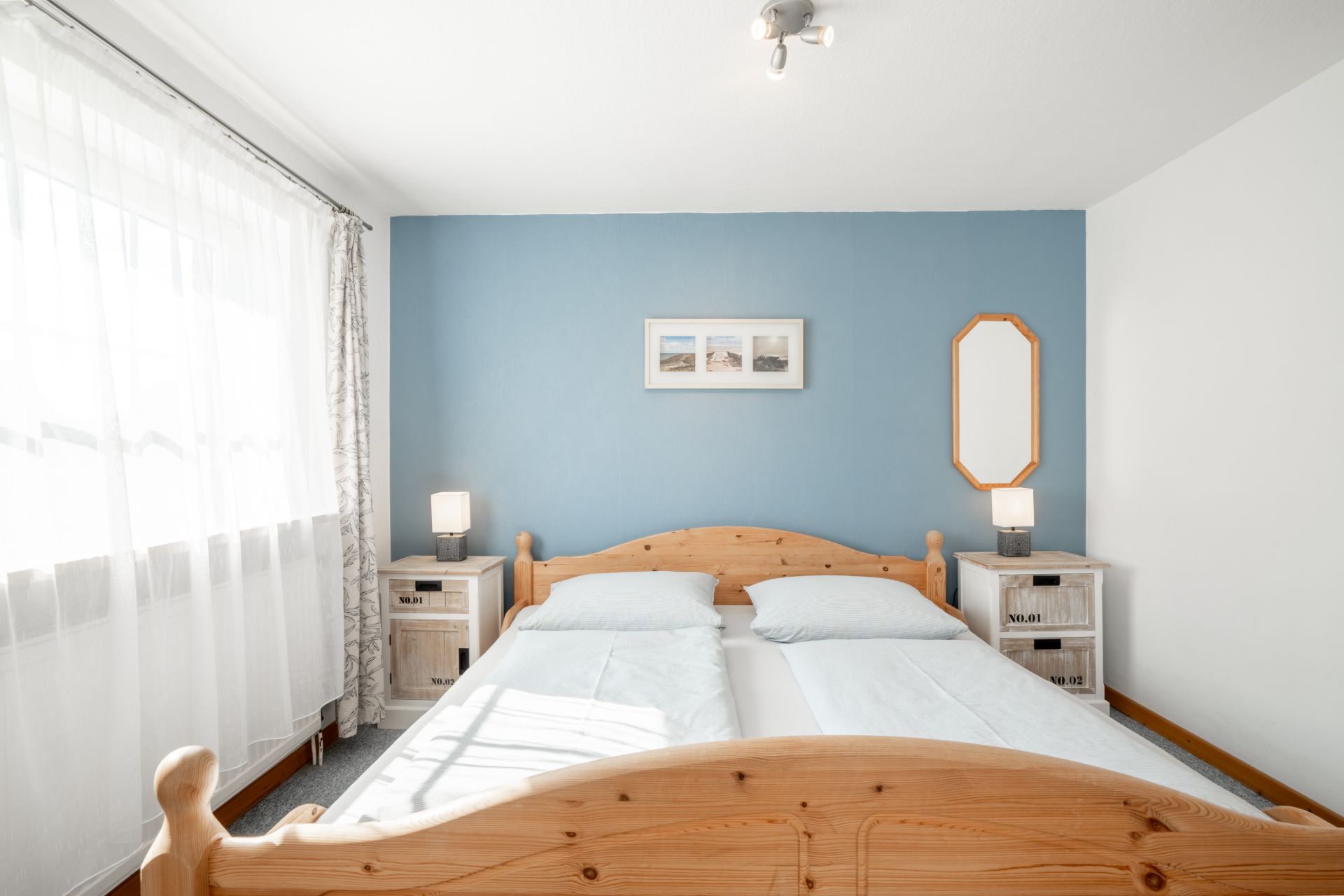 Helles Schlafzimmer mit Doppelbett, blauer Akzentwand, Nachttischen und großem Fenster mit Vorhängen.