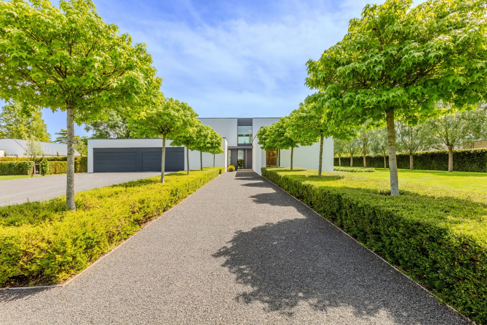 Moderner Eingangsweg zu einem weißen Wohnhaus mit gepflegtem Garten und Bäumen.