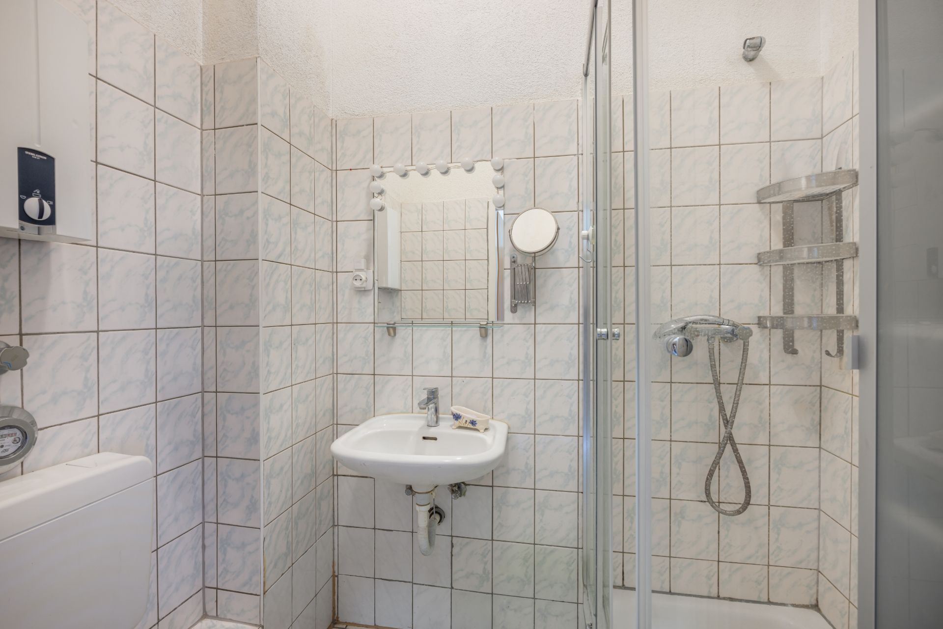 Kleines Badezimmer mit Waschbecken, Spiegel und Dusche mit Handbrause, alles gefliest.