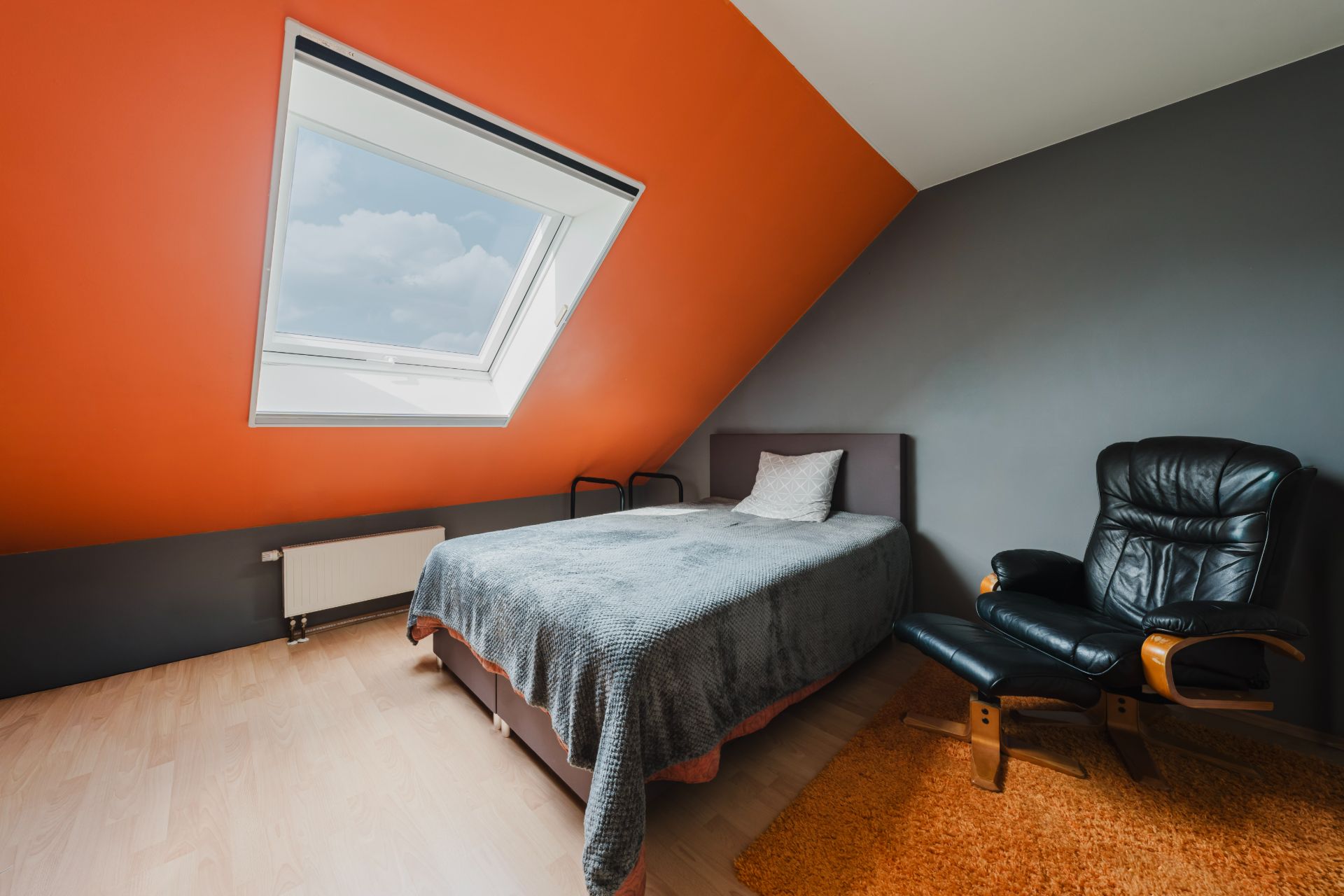Dachzimmer mit Bett, Relaxsessel und schrägem Fenster in orange-grauer Wand.