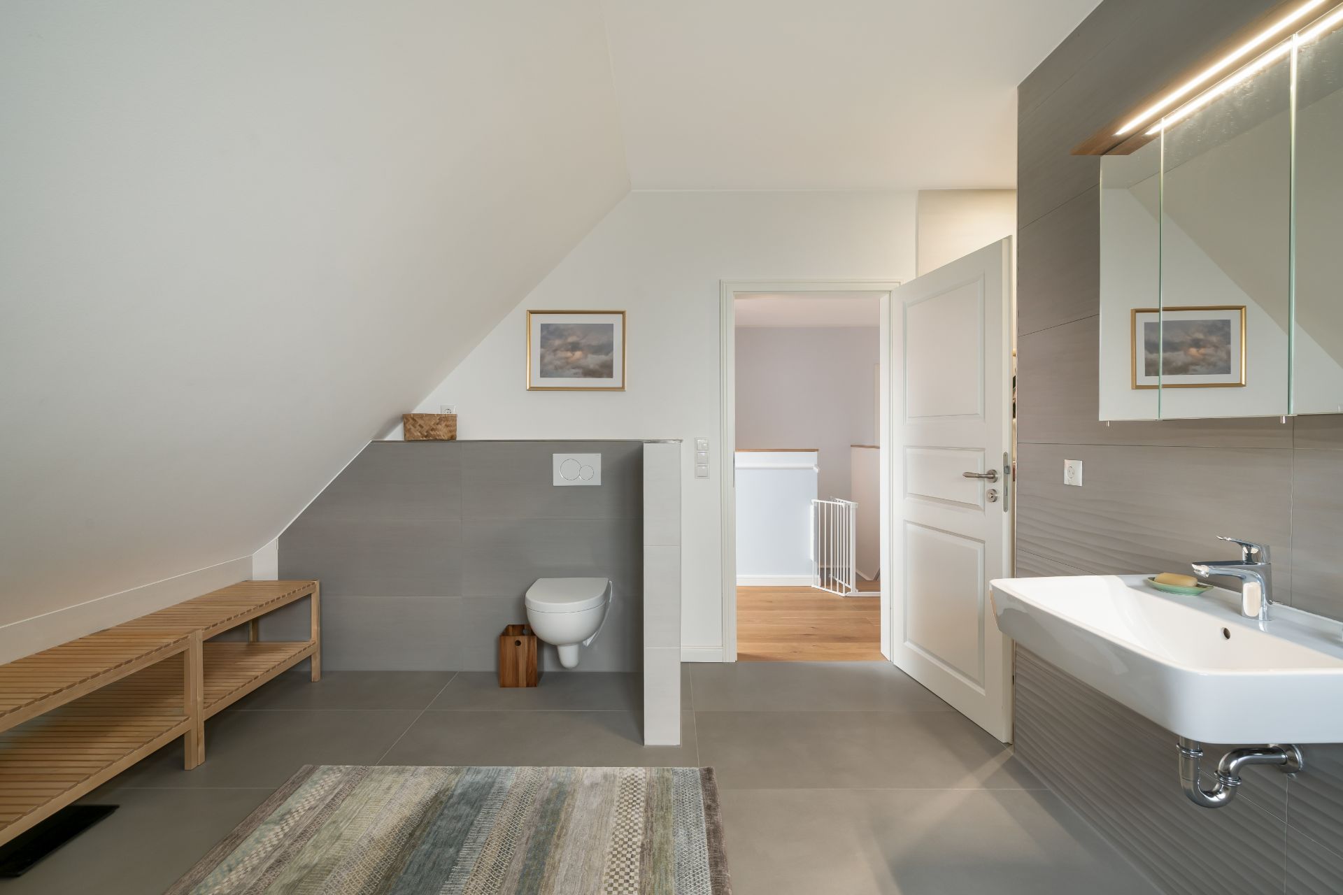 Modernes Badezimmer mit Wandtoilette, Waschbecken, Spiegel und Holzbank unter Dachschräge.