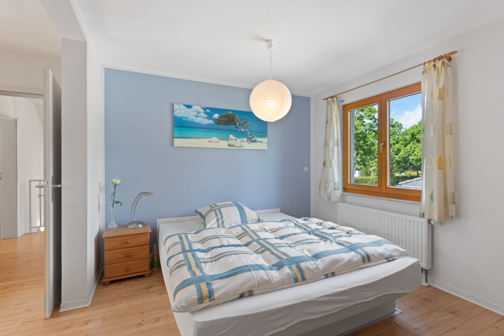Helles Schlafzimmer mit Doppelbett, Nachttisch, Fenster und Bild an blauer Wand.