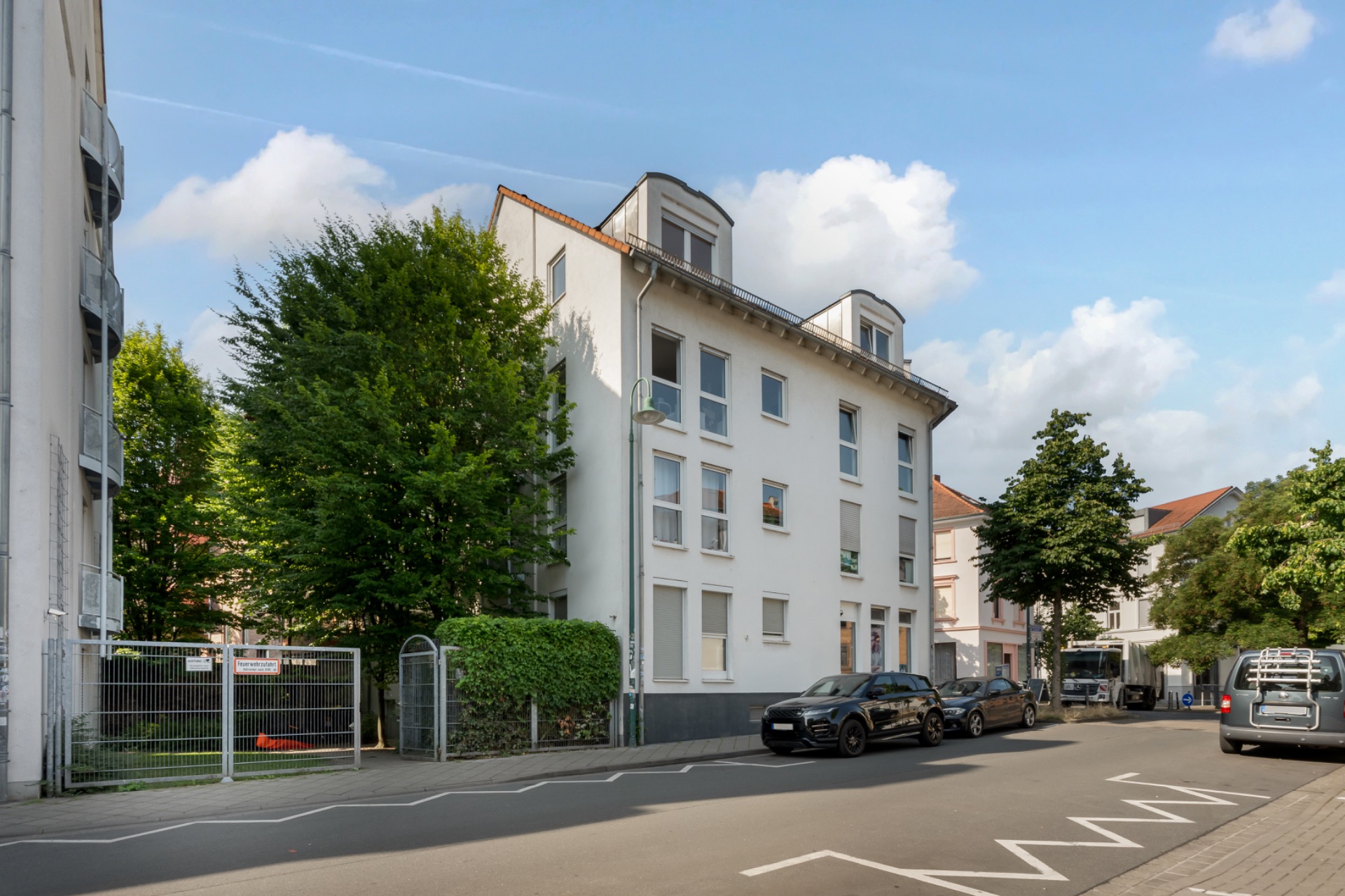 Wohnhaus mit drei Stockwerken an einer ruhigen Straße, mehrere Autos parken am Straßenrand.
