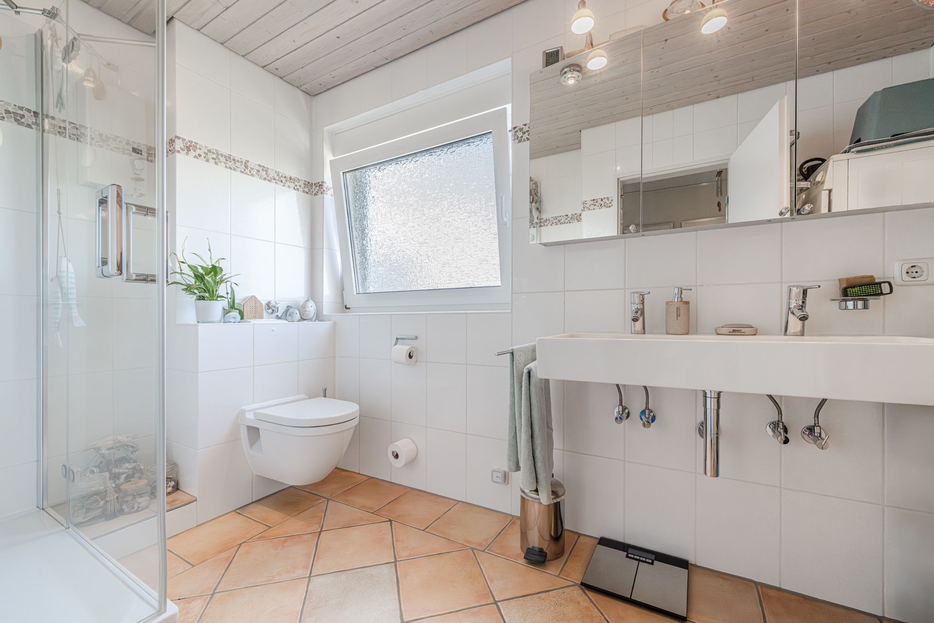 Modernes, helles Badezimmer mit Dusche, Toilette, Doppelwaschbecken und großem Fenster.
