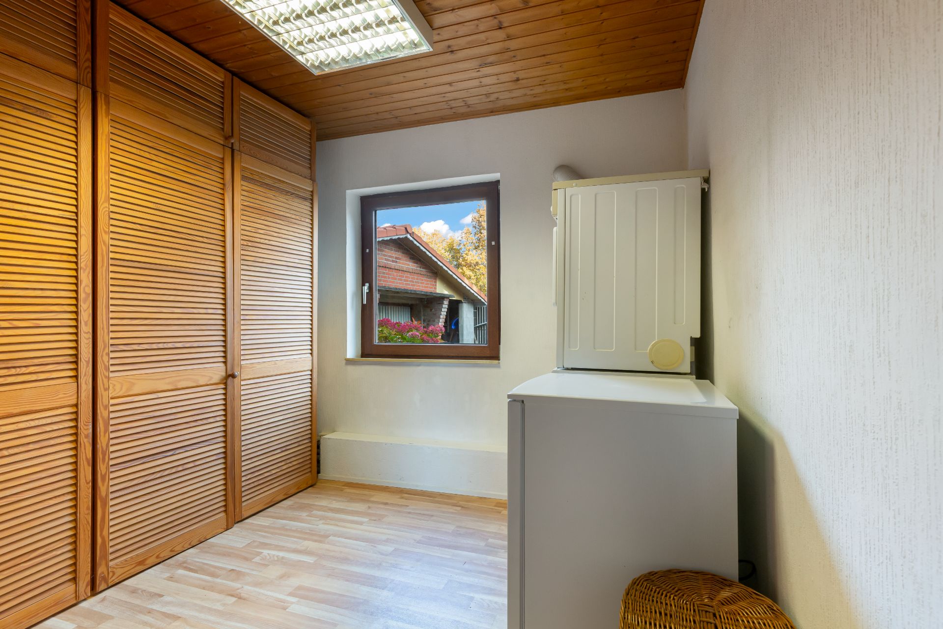 Kleiner Raum mit Holzschränken, Fenster, weißem Schrank und Korb auf Laminatboden.