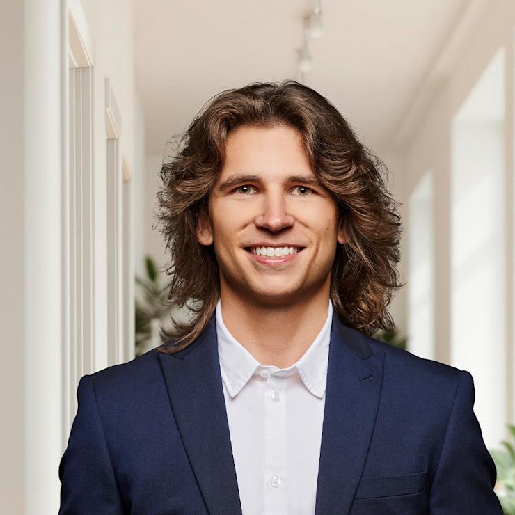 Junger Mann im Anzug steht in einem hellen Flur mit Pflanzen im Hintergrund.