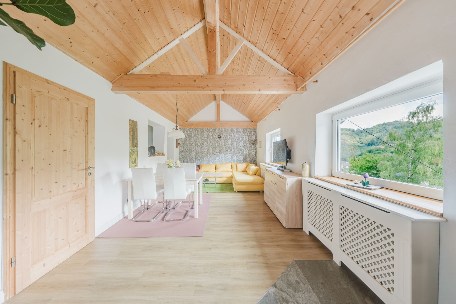 Helles Wohnzimmer mit Holzdecke, gelbem Sofa, Esstisch und großem Fenster mit Ausblick.