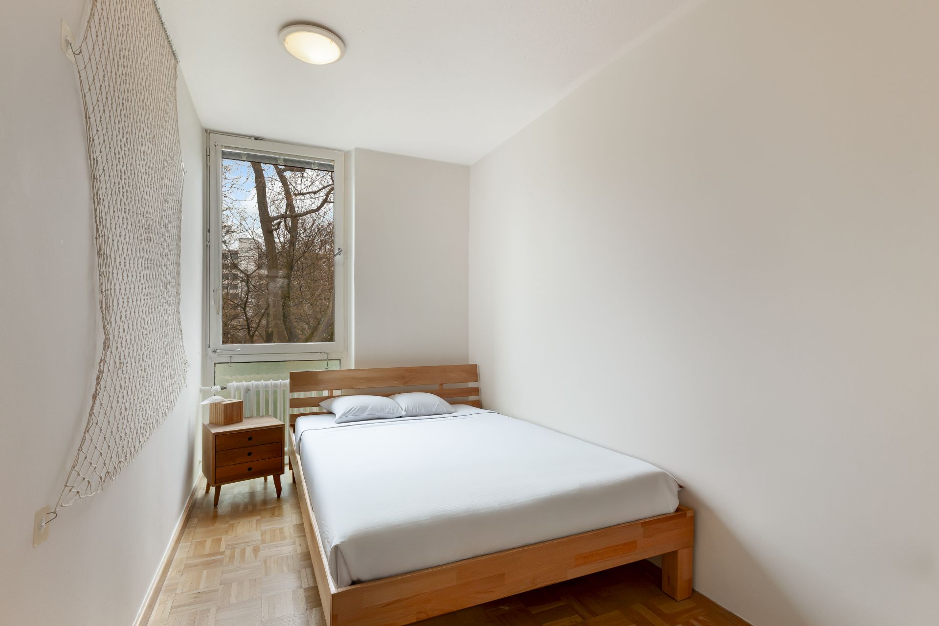 Helles Schlafzimmer mit großem Bett, Nachttisch und Fenster mit Blick auf Bäume.