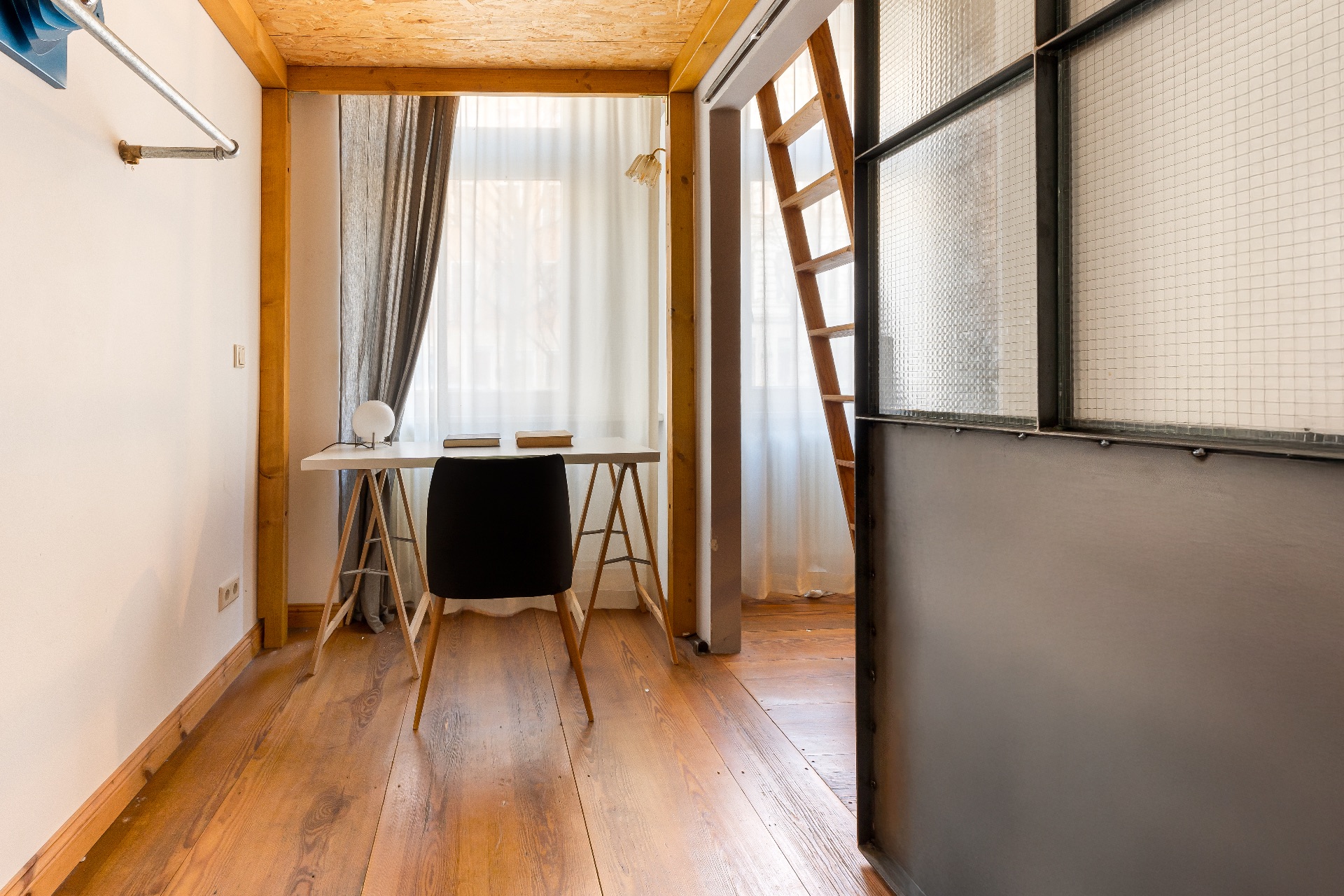 Helles Zimmer mit Schreibtisch, Stuhl, Holzfußboden und einer Leiter, die zu einer oberen Etage führt.