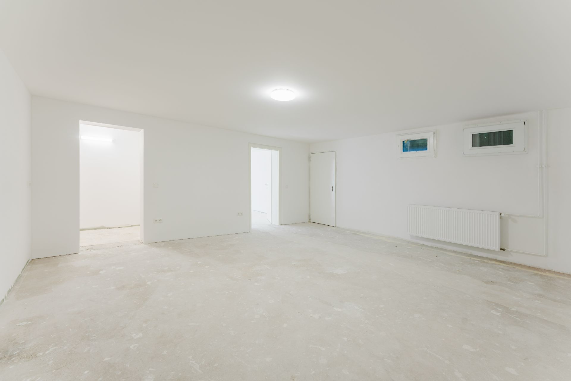 Leerer, weiß gestrichener Raum mit zwei Türen, kleinen Fenstern und einem Heizkörper an der Wand.