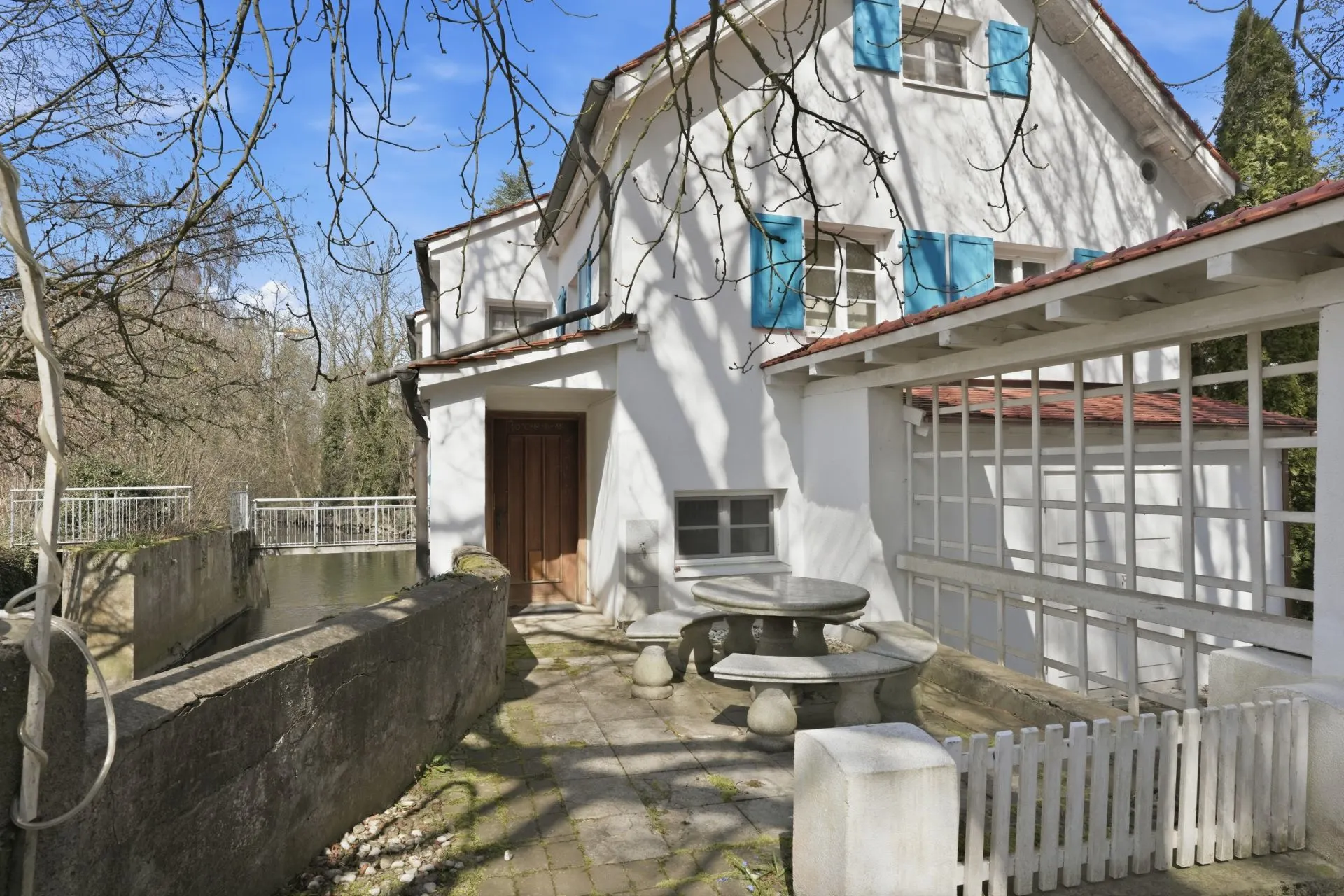 Weißes Haus mit blauen Fensterläden, Steintisch und Bänken auf einer Terrasse.