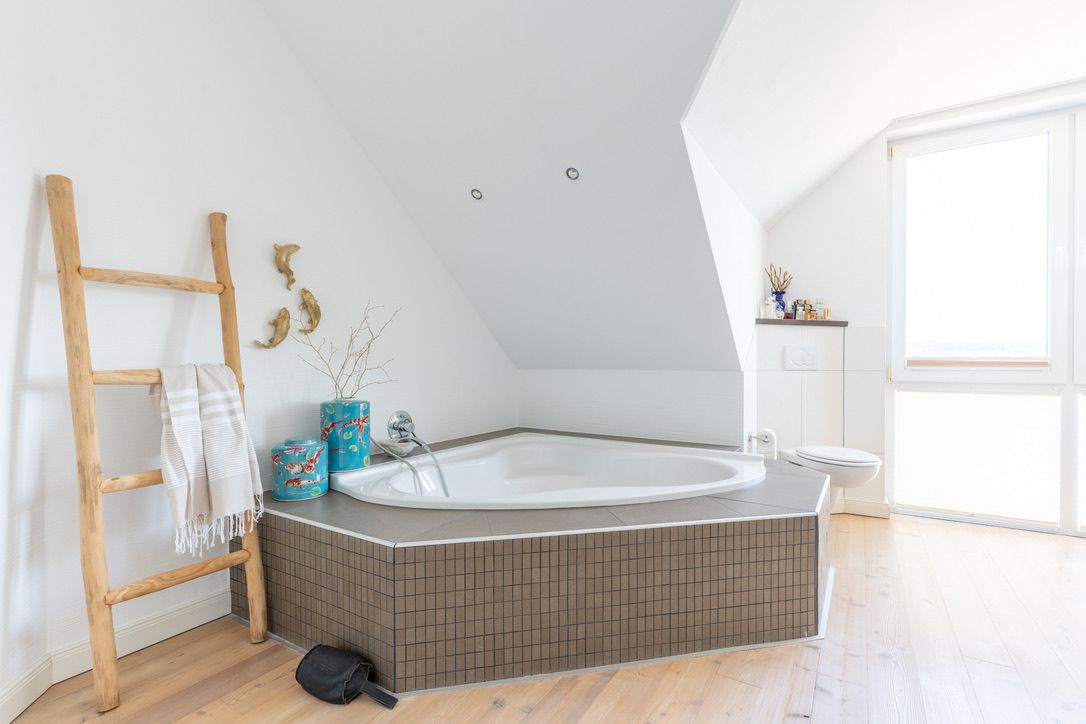 Helles Badezimmer mit Eckbadewanne, Holzleiter und großem Fenster.