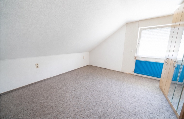 Leeres Zimmer mit Dachschräge, Teppichboden, Fenster mit blauem Heizkörper und Spiegelschrank.